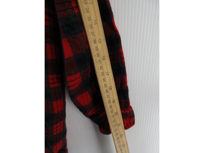 Puede incluir: Una camisa de franela de manga larga con un patr&oacute;n de cuadros rojos y negros. Una regla de madera est&aacute; colocada verticalmente a lo largo de la manga, mostrando medidas de aproximadamente 33 cm a 56 cm para indicar la longitud.