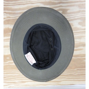 Op de afbeelding: Een olijfgroene hoed met een brede rand en een donker interieur. De hoed heeft een gestructureerde rand en een zwarte voering. Een wit label is zichtbaar aan de binnenkant. De hoed is waarschijnlijk gemaakt van een duurzame stof, geschikt voor buiten.