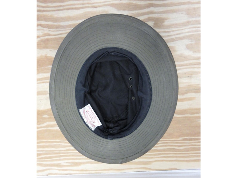 Op de afbeelding: Een olijfgroene hoed met een brede rand en een donker interieur. De hoed heeft een gestructureerde rand en een zwarte voering. Een wit label is zichtbaar aan de binnenkant. De hoed is waarschijnlijk gemaakt van een duurzame stof, geschikt voor buiten.