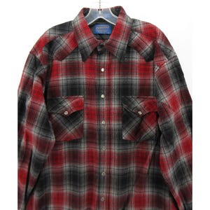 May include: A long-sleeved button-down shirt featuring a red, black and grey plaid pattern. The shirt includes a pointed collar, two chest pockets with button-down flaps, and silver-toned snap buttons down the front placket. A Pendleton label is visible.