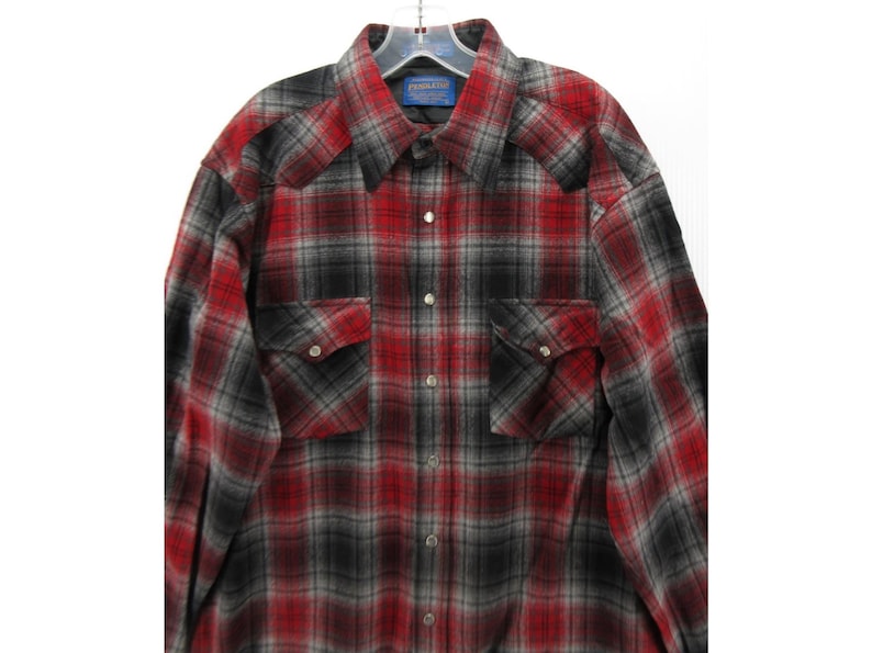May include: A long-sleeved button-down shirt featuring a red, black and grey plaid pattern. The shirt includes a pointed collar, two chest pockets with button-down flaps, and silver-toned snap buttons down the front placket. A Pendleton label is visible.