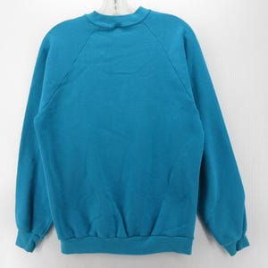May include: A solid teal long-sleeve sweatshirt featuring a crew neckline, raglan sleeves, and ribbed cuffs at the wrists and hem. The garment is a plain, unpatterned pullover style with a simple construction and a consistent color throughout.