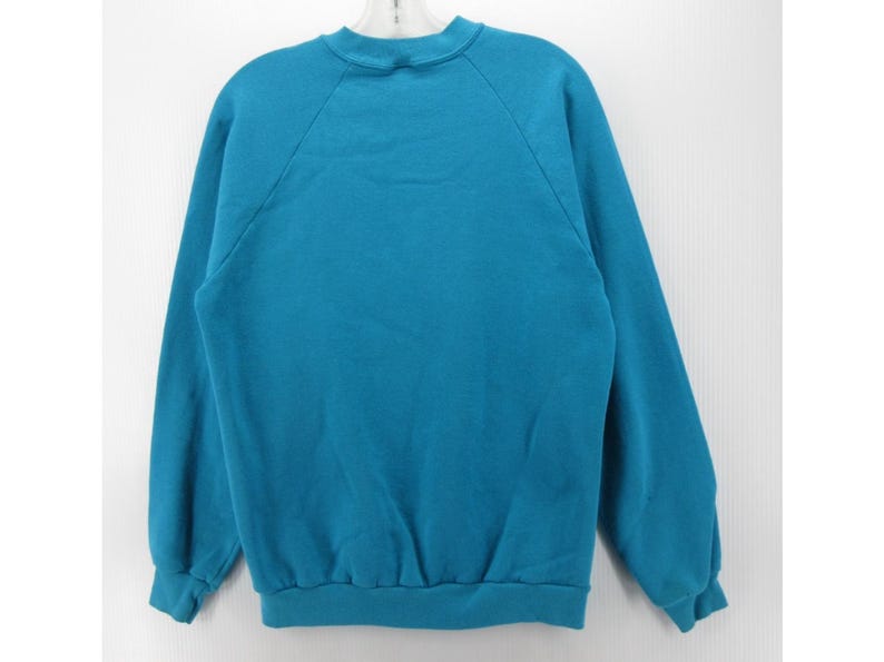 May include: A solid teal long-sleeve sweatshirt featuring a crew neckline, raglan sleeves, and ribbed cuffs at the wrists and hem. The garment is a plain, unpatterned pullover style with a simple construction and a consistent color throughout.