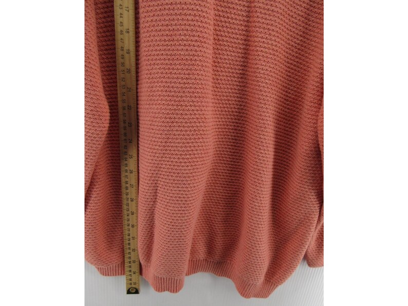 May include: A coral-colored sweater with a textured knit pattern. The sweater has a ribbed hem and cuffs. A wooden ruler is placed vertically along the side of the sweater, showing measurements in inches.