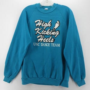 May include: A teal long-sleeve crewneck sweatshirt featuring white text that reads High Kicking Heels with a small white footprint graphic above the text and the words UNC DANCE TEAM printed in white below the main graphic on the chest.
