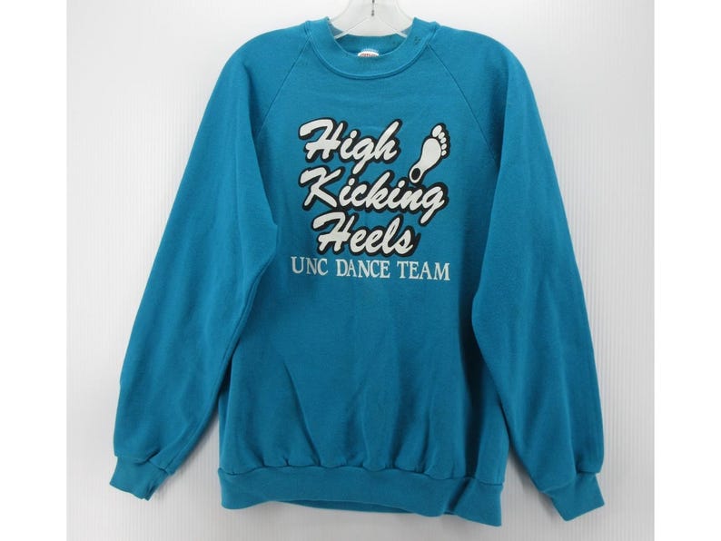 May include: A teal long-sleeve crewneck sweatshirt featuring white text that reads High Kicking Heels with a small white footprint graphic above the text and the words UNC DANCE TEAM printed in white below the main graphic on the chest.