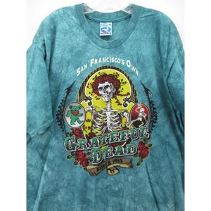 May include: A turquoise tie-dye t-shirt featuring a skeleton with a red afro and a cigarette, surrounded by roses and the words "Grateful Dead" and "San Francisco's Own". The shirt also has a dancing bear graphic.