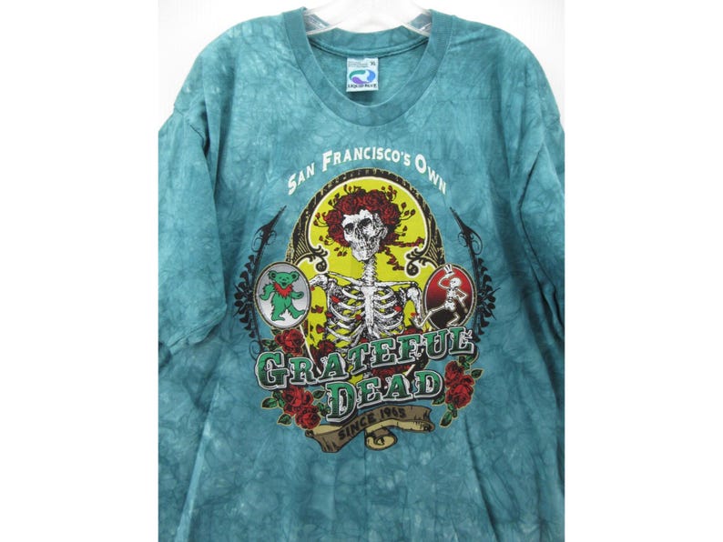 May include: A turquoise tie-dye t-shirt featuring a skeleton with a red afro and a cigarette, surrounded by roses and the words "Grateful Dead" and "San Francisco's Own". The shirt also has a dancing bear graphic.