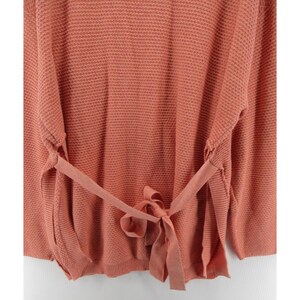 May include: A coral-colored sweater with a textured knit pattern. The sweater features long sleeves and a tie-waist detail, creating a flattering silhouette. The tie is made of the same material as the sweater. The overall style is casual and comfortable.
