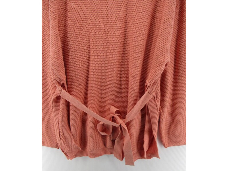 May include: A coral-colored sweater with a textured knit pattern. The sweater features long sleeves and a tie-waist detail, creating a flattering silhouette. The tie is made of the same material as the sweater. The overall style is casual and comfortable.