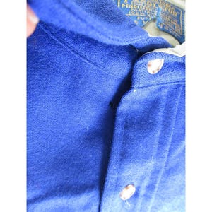 May include: A royal blue polo shirt with a textured fabric finish, featuring a button-up placket with two visible light-colored buttons. A Pendleton brand label is stitched inside the collar, displaying text including Portland and 100% wool.