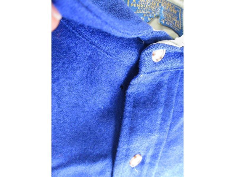 May include: A royal blue polo shirt with a textured fabric finish, featuring a button-up placket with two visible light-colored buttons. A Pendleton brand label is stitched inside the collar, displaying text including Portland and 100% wool.