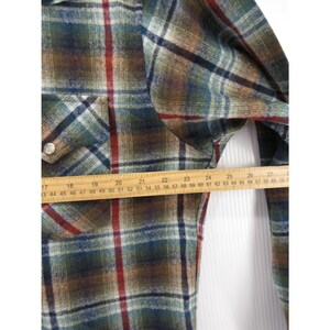 May include: A long-sleeve plaid shirt featuring a pattern of green, brown, blue, and red lines. A wooden ruler is placed horizontally across the chest, showing a measurement spanning from approximately 43 cm to 69 cm in width.