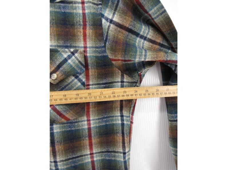 May include: A long-sleeve plaid shirt featuring a pattern of green, brown, blue, and red lines. A wooden ruler is placed horizontally across the chest, showing a measurement spanning from approximately 43 cm to 69 cm in width.