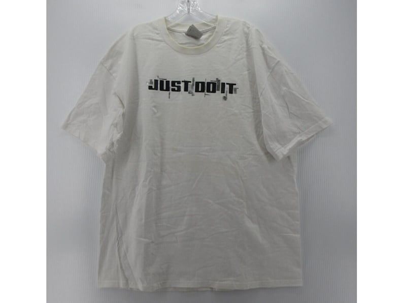 May include: A white short-sleeved t-shirt with the text "JUST DO IT" in black, with a gray Nike logo on the upper chest. The shirt is hanging on a clear hanger against a white background.