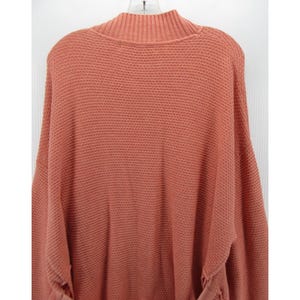 May include: A coral-colored sweater with a mock neck and long sleeves. The sweater has a textured knit pattern and is hanging on a white hanger. The sleeves have a gathered detail at the cuffs. The sweater is a solid color.