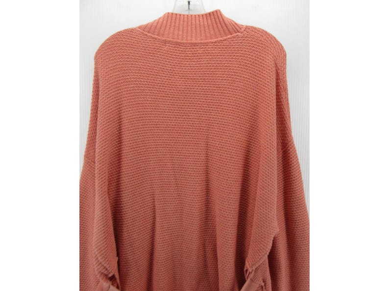 May include: A coral-colored sweater with a mock neck and long sleeves. The sweater has a textured knit pattern and is hanging on a white hanger. The sleeves have a gathered detail at the cuffs. The sweater is a solid color.