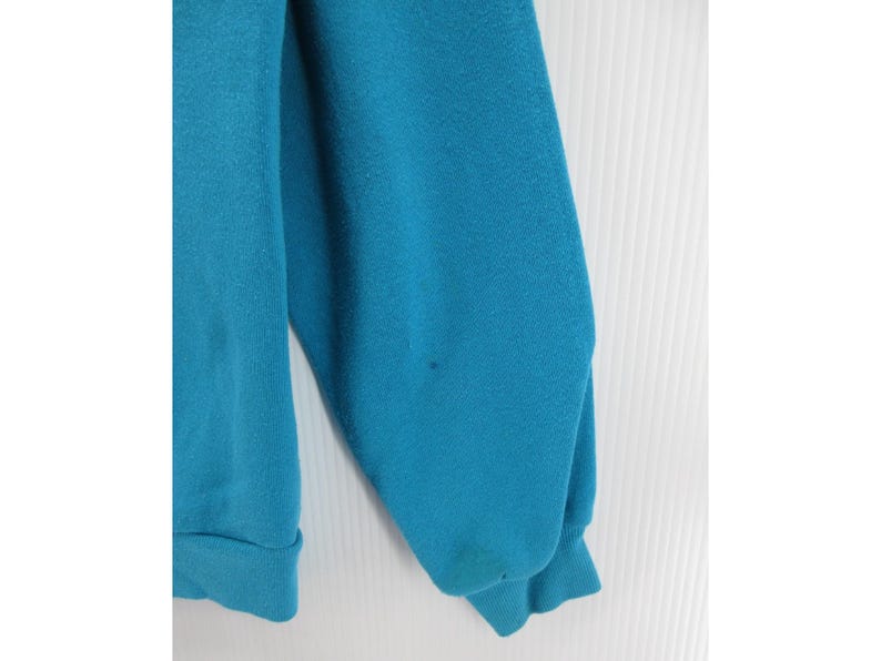 May include: A solid teal long-sleeve sweatshirt featuring a crew neckline and ribbed cuffs at the wrists. The garment is constructed from a soft, fleece-like fabric and displays a plain, unpatterned design with a consistent vibrant blue-green color.
