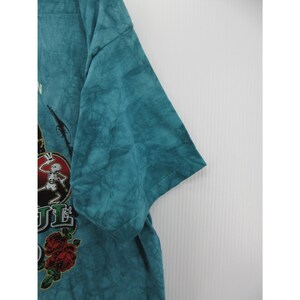 May include: Teal tie-dye t-shirt featuring a graphic of a skeleton in a hat, roses, and the text "Grateful Dead". The shirt has short sleeves and a crew neck. The fabric has a textured appearance.