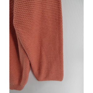 May include: Close-up of a coral-colored sweater sleeve. The knitwear has a textured, horizontal ribbing pattern. The sleeve is folded, showing the cuff and part of the body of the sweater. The background is white.
