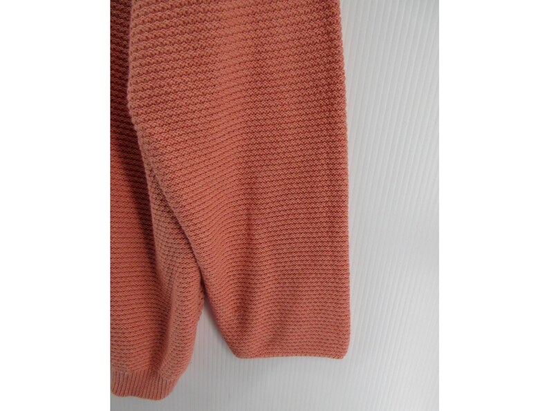 May include: Close-up of a coral-colored sweater sleeve. The knitwear has a textured, horizontal ribbing pattern. The sleeve is folded, showing the cuff and part of the body of the sweater. The background is white.