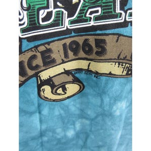 May include: Close-up of a blue tie-dye t-shirt with a graphic design. The design includes the text "ICE" in a bold, green font, and a banner that reads "SINCE 1965". The shirt has a vintage aesthetic.