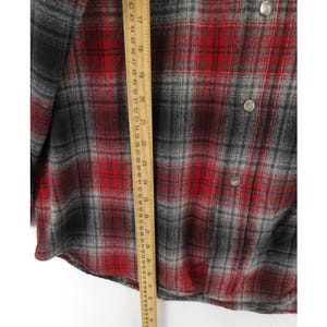 May include: A long-sleeve button-down shirt featuring a red, black, and grey plaid pattern. A wooden ruler is placed vertically over the fabric, showing a length measurement extending from approximately 41 cm to 74 cm on the metric scale.