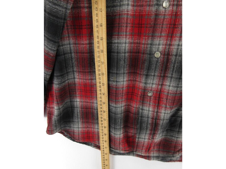 May include: A long-sleeve button-down shirt featuring a red, black, and grey plaid pattern. A wooden ruler is placed vertically over the fabric, showing a length measurement extending from approximately 41 cm to 74 cm on the metric scale.