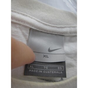 May include: Close-up of a white t-shirt collar with a tag. The tag features the Nike logo and the size XL. The text "MADE IN GUATEMALA" is printed on the tag.