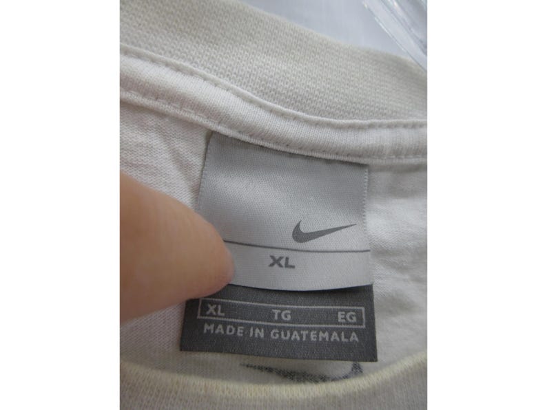 May include: Close-up of a white t-shirt collar with a tag. The tag features the Nike logo and the size XL. The text "MADE IN GUATEMALA" is printed on the tag.