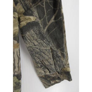 Puede incluir: Una camisa de manga larga con un estampado de camuflaje en tonos marrones, verdes y grises. La tela tiene un estampado detallado que se asemeja a un entorno forestal. El pu&ntilde;o de la manga est&aacute; abotonado.
