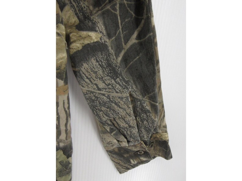 Puede incluir: Una camisa de manga larga con un estampado de camuflaje en tonos marrones, verdes y grises. La tela tiene un estampado detallado que se asemeja a un entorno forestal. El pu&ntilde;o de la manga est&aacute; abotonado.