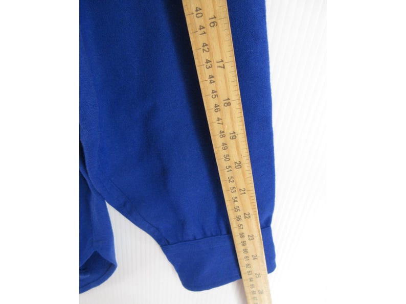 May include: A solid royal blue long-sleeved shirt with a buttoned cuff. A wooden ruler is placed vertically along the sleeve, showing a measurement of approximately 25 inches from the shoulder seam to the end of the cuff.