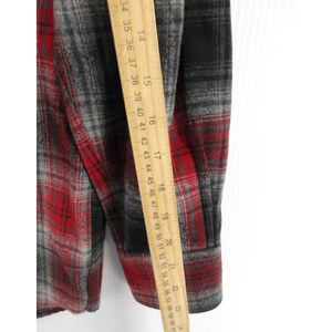 May include: A long-sleeve flannel shirt featuring a red, black, and grey plaid pattern. A wooden ruler is placed vertically against the fabric, showing measurements from 35 cm to 58 cm along the sleeve and side of the garment.