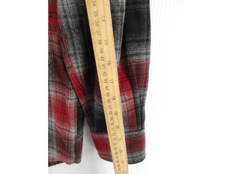 May include: A long-sleeve flannel shirt featuring a red, black, and grey plaid pattern. A wooden ruler is placed vertically against the fabric, showing measurements from 35 cm to 58 cm along the sleeve and side of the garment.