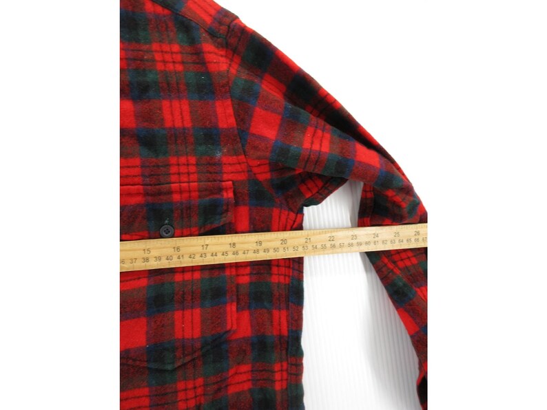 Puede incluir: Una camisa de franela de manga larga con un patr&oacute;n de cuadros en rojo, negro y verde oscuro, que incluye un bolsillo en el pecho con bot&oacute;n. Una regla de madera colocada sobre la parte central muestra una medida de unos 28 cent&iacute;metros.