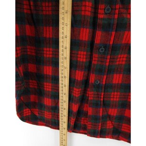 Puede incluir: Una camisa de franela a cuadros rojos y verde oscuro con cierre de botones. Una regla de madera est&aacute; colocada verticalmente sobre la tela, mostrando una medida de longitud que va desde aproximadamente 38 cm a 71 cm en la escala m&eacute;trica.