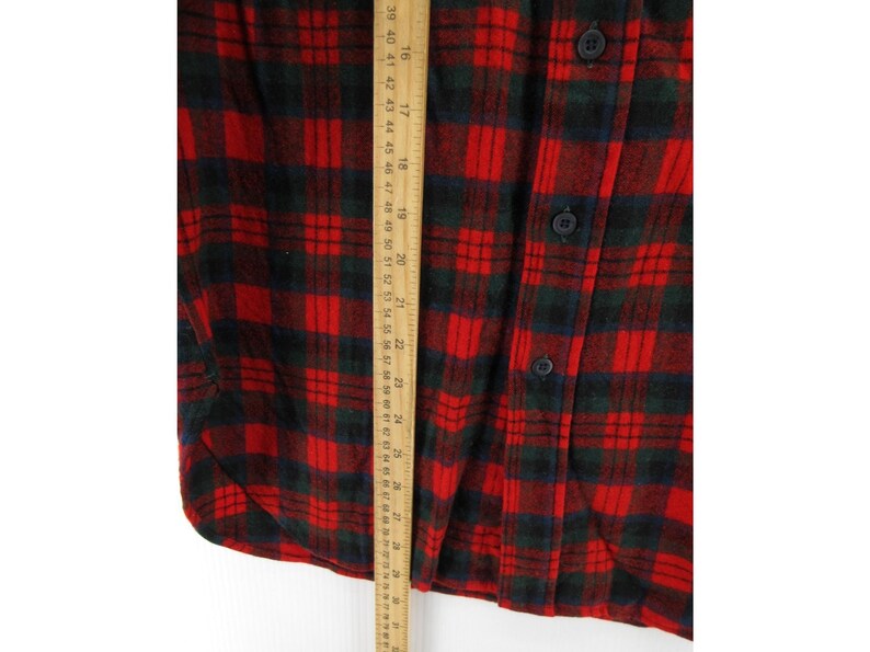 Puede incluir: Una camisa de franela a cuadros rojos y verde oscuro con cierre de botones. Una regla de madera est&aacute; colocada verticalmente sobre la tela, mostrando una medida de longitud que va desde aproximadamente 38 cm a 71 cm en la escala m&eacute;trica.