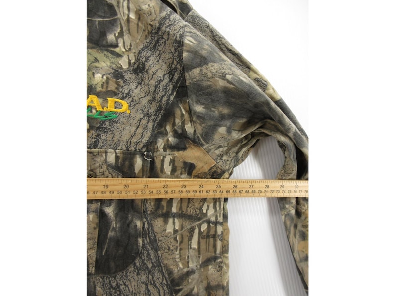 Puede incluir: Chaqueta con estampado de camuflaje con el texto "A.D." en bordado amarillo y verde. La chaqueta tiene una cremallera y una regla colocada en la parte delantera, que mide en cent&iacute;metros.