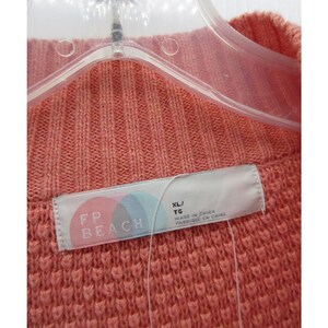 May include: Close-up of a coral-colored knit sweater. The sweater is hanging on a clear plastic hanger. A tag with the text "FP BEACH" and size "XL/TG" is visible. The sweater has a textured, ribbed design.