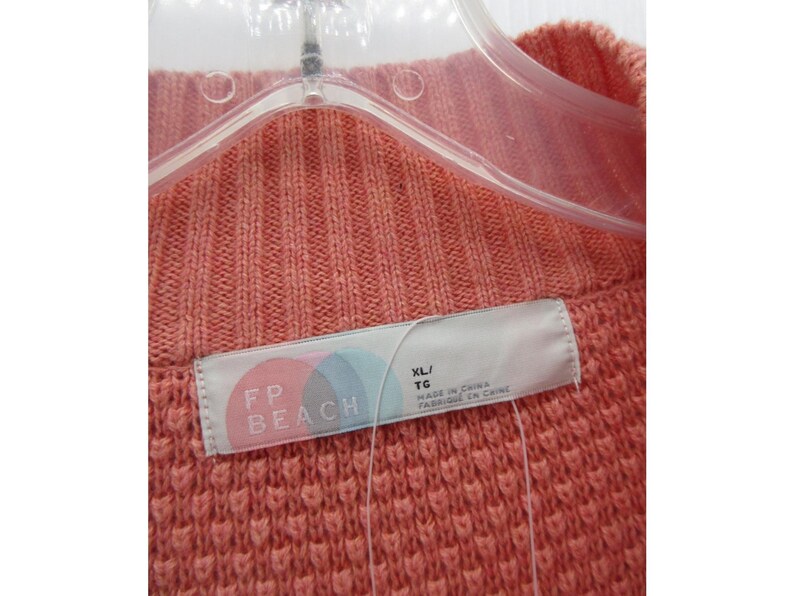 May include: Close-up of a coral-colored knit sweater. The sweater is hanging on a clear plastic hanger. A tag with the text "FP BEACH" and size "XL/TG" is visible. The sweater has a textured, ribbed design.