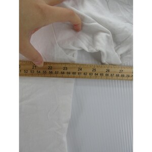 May include: A white garment is being measured with a wooden ruler. The ruler shows measurements from 21 to 27 inches. The fabric is crumpled and appears to be made of a soft material. The background is a white, ridged surface.