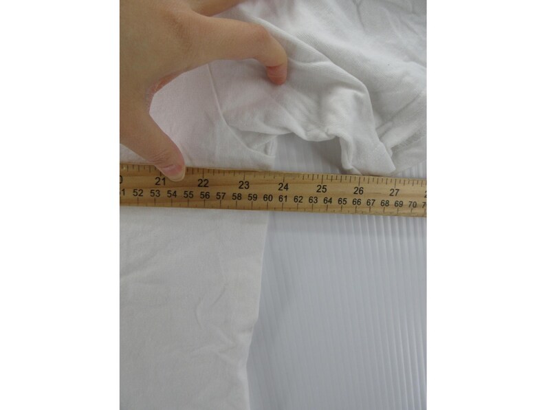 May include: A white garment is being measured with a wooden ruler. The ruler shows measurements from 21 to 27 inches. The fabric is crumpled and appears to be made of a soft material. The background is a white, ridged surface.