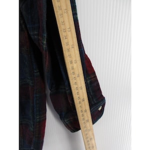 May include: A long-sleeved flannel shirt featuring a dark plaid pattern in shades of navy blue, forest green, and deep red. A wooden ruler is placed vertically along the sleeve, showing measurements from 12 inches to 22 inches in length.