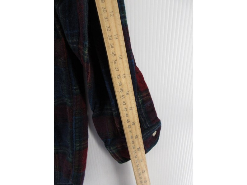 May include: A long-sleeved flannel shirt featuring a dark plaid pattern in shades of navy blue, forest green, and deep red. A wooden ruler is placed vertically along the sleeve, showing measurements from 12 inches to 22 inches in length.