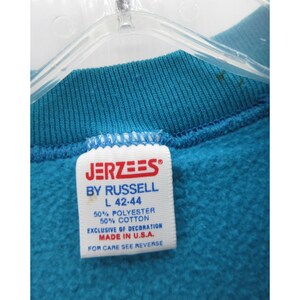 May include: A teal sweatshirt featuring a white interior tag with red and blue text that reads JERZEES BY RUSSELL, size L 42-44, 50% polyester, 50% cotton, exclusive of decoration, made in U.S.A., and for care see reverse.