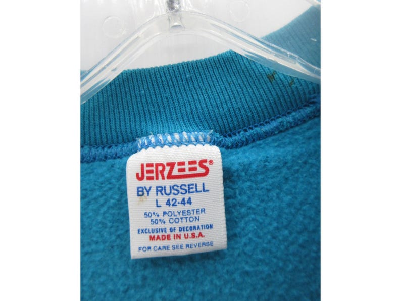 May include: A teal sweatshirt featuring a white interior tag with red and blue text that reads JERZEES BY RUSSELL, size L 42-44, 50% polyester, 50% cotton, exclusive of decoration, made in U.S.A., and for care see reverse.