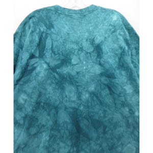May include: A teal tie-dye t-shirt with a crew neck. The fabric has a textured, marbled appearance, with darker teal shades creating a pattern across the shirt. The shirt is hanging on a white hanger.