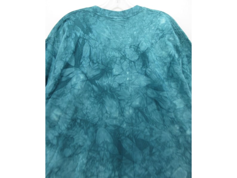 May include: A teal tie-dye t-shirt with a crew neck. The fabric has a textured, marbled appearance, with darker teal shades creating a pattern across the shirt. The shirt is hanging on a white hanger.