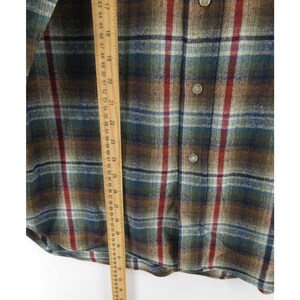 May include: A long-sleeve button-down shirt featuring a plaid pattern in shades of brown, dark green, blue, and red. A wooden ruler is placed vertically over the fabric, showing measurements in inches and centimetres for scale and reference.